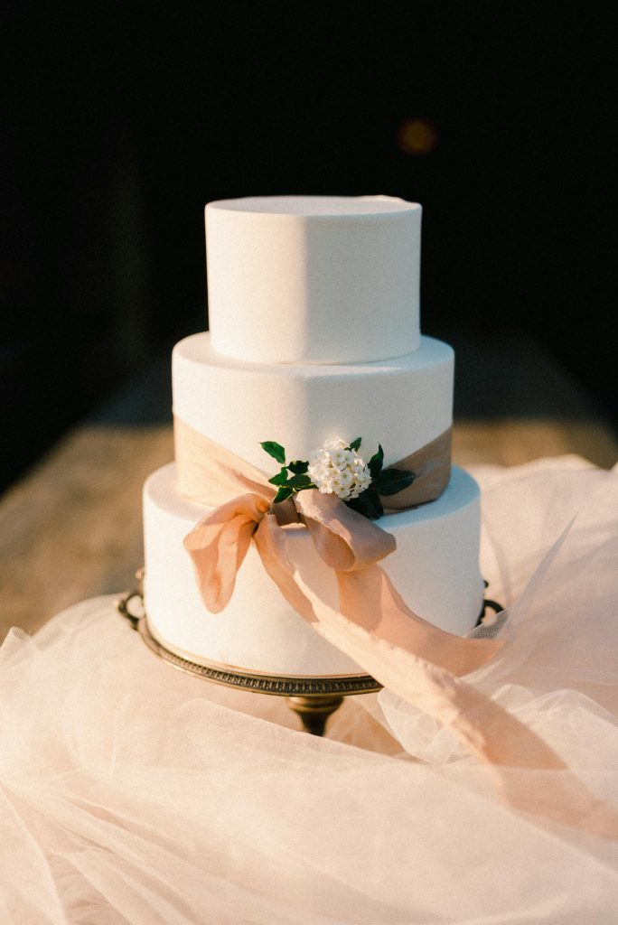 A classic three-tier wedding cake adorned with a beige ribbon and white flowers on a stand.